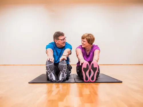 Kaksi henkilöä venyttelee jumppamatolla sisätiloissa, hymyilevät toisilleen.