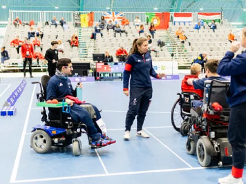 Hallissa peli pyörätuolissa, pelaajat ja valmentaja keskustelevat. Taustalla katsojia.