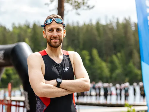 Mies triathlon-puvussa seisoo kädet ristissä, taustalla järvimaisema ja banneri.