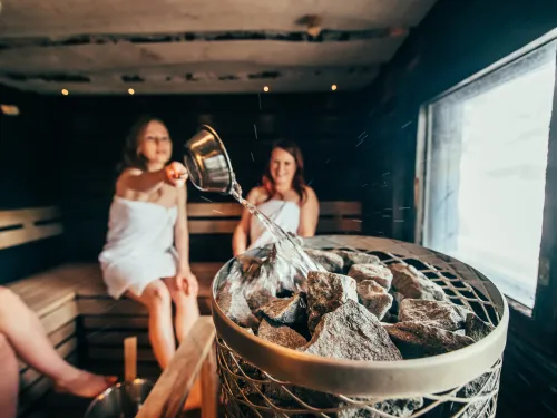 Naiset istuvat saunan lauteilla ja yksi heittää löylyä kiukaalle.