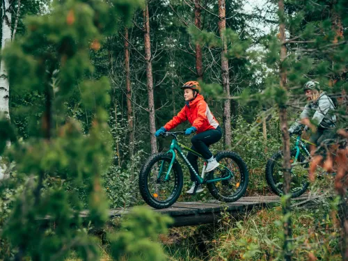 Kaksi ihmistä pyöräilee fatbikeilla metsässä ojan yli menevän sillan päällä.