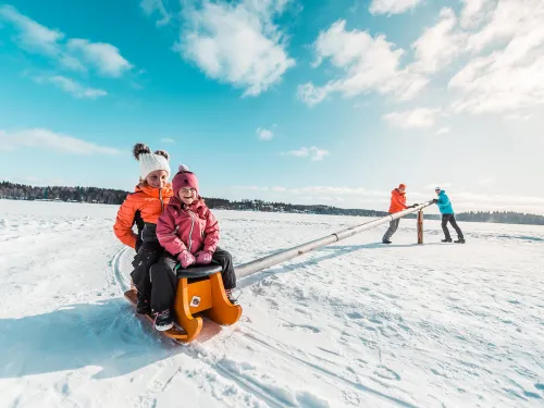 Kaksi lasta talvivaatteissa jäällä napakelkassa järven jäällä aikuisten työntäessä.