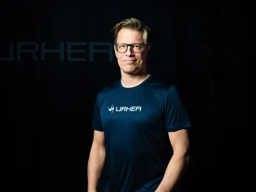 Henkilö seisoo tummassa ympäristössä, päällään sininen paita ja silmälasit.