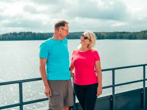 Man and woman smiling at each other on a boat, lake and trees in background.