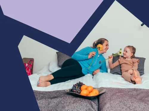 Nainen ja tyttö makoilevat sängyllä toisiaan katsellen kukat kädessään.