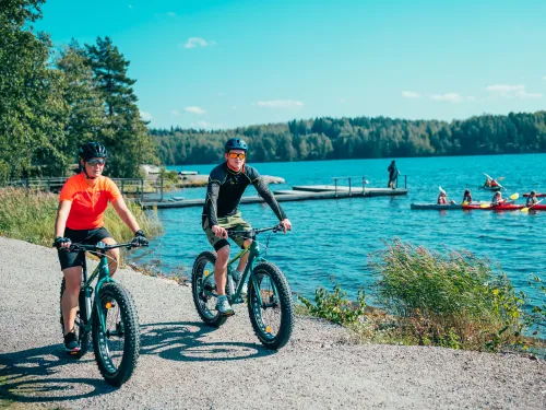 Mies ja nainen pyöräilevät fatbikeillä järven rannalla ja taustalla ihmisiä melomassa.