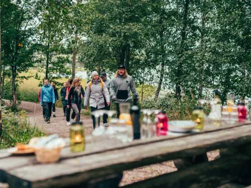 Polulla kävelee useita ihmisiä metsässä. Etualalla on katettu puupöytä tuikkuineen..