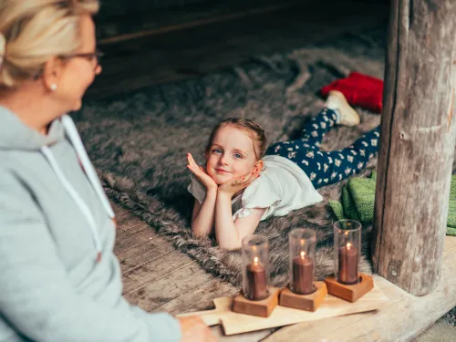 Hymyilevä tyttö makaa laavulla taljan päällä mummon katsoessa vieressä.