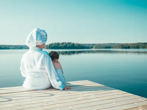 Nainen valkoisessa kylpytakissa ja tyttö pyyhe ympärillään istuvat laiturilla katsellen tyyntä, kesäistä järveä.