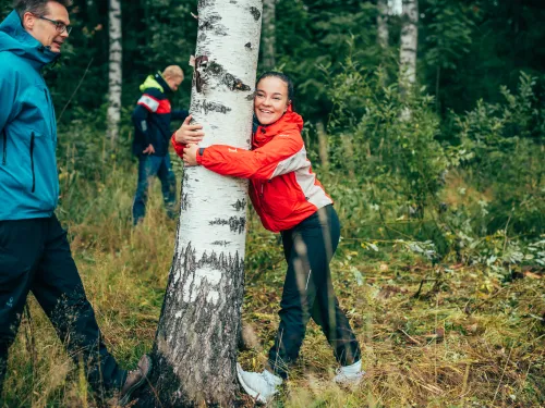 Nainen venyttelee jalkaa puuta vasten, hymyilee ja halaa puun runkoa
