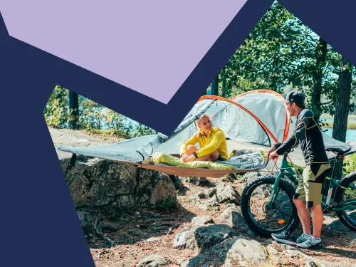 Järven rannalla puihin on kiinnitetty tentsile, jossa nainen istuu ja hymyilee fatbikea pitelevälle miehelle.