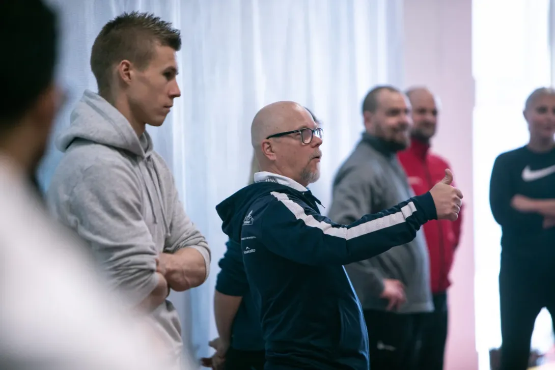 Useat miehet seisovat keskustellen sisätiloissa, yksi elehtii kädellään.