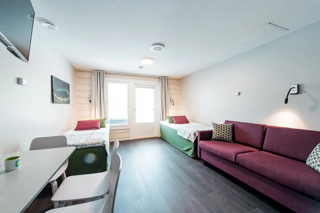 Valoisa hotellihuone, kaksi sänkyä, sohvapöytä, violetti sohva.