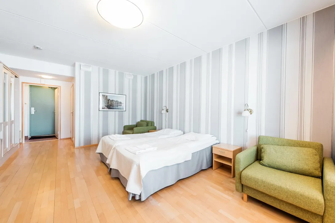 Valoisa hotellihuone, kaksi sänkyä ja vaaleanvihreät nojatuolit.