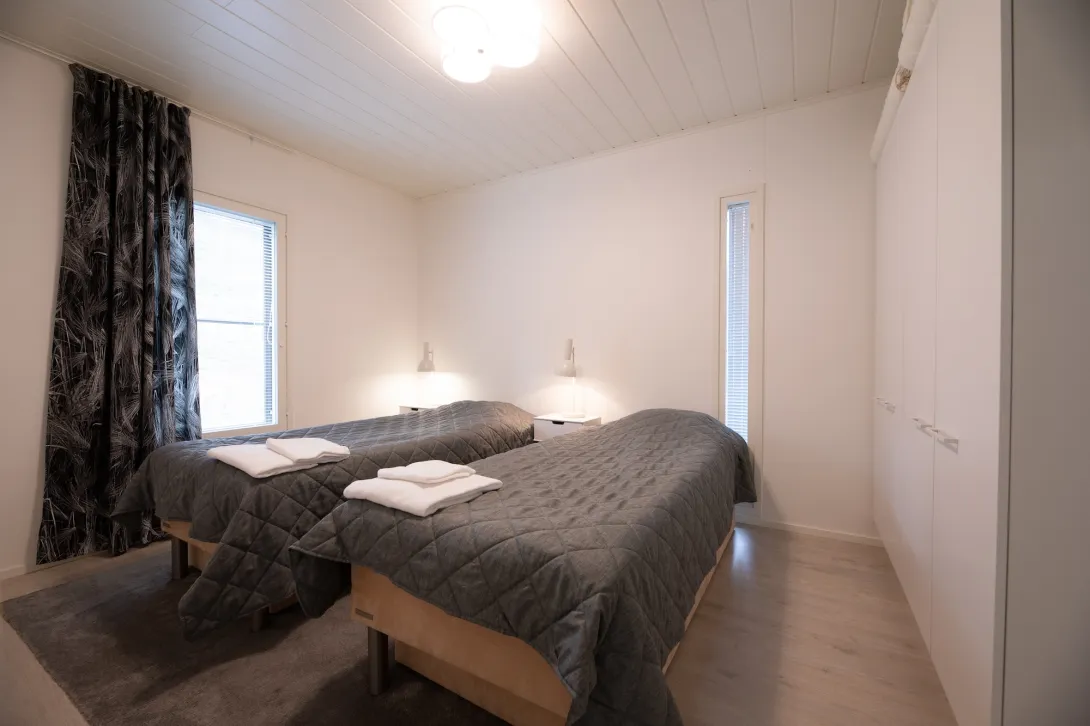 Kaksi sänkyä valkoisessa makuuhuoneessa, harmaat peitot, ikkunat vasemmalla.