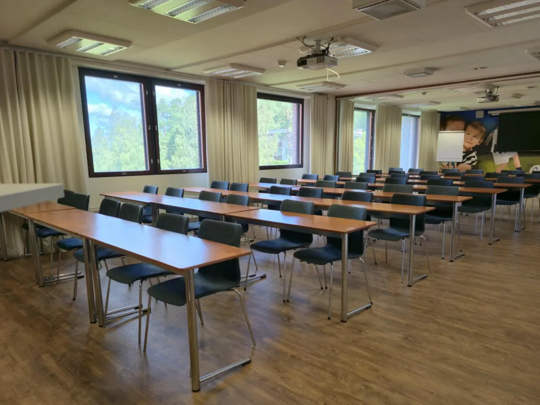 Tyhjä luokkahuone, puiset pöydät, mustat tuolit, ikkunat taustalla.